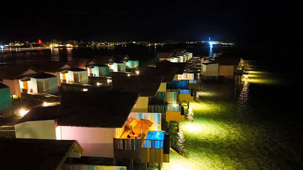 Rij felverlichte watervilla's 's nachts met gloeiend water eronder bij Kurumba Maldives resort.