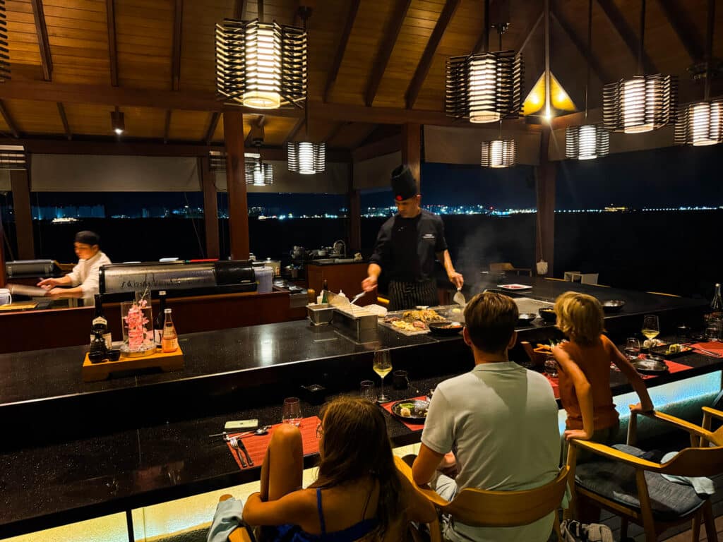 Gezin geniet van een live teppanyaki-dinershow met stadslichten op de achtergrond bij Kurumba Maldives resort.