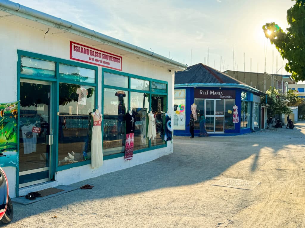 Small souvenir shops in Dharavandhoo Maldives displaying dresses and beachwear along a sandy street near the harbor at sunset.