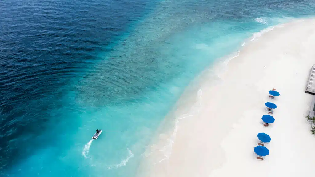Los mejores resorts económicos en Maldivas: 10 opciones asequibles 13 vista aérea de una moto de agua cruzando aguas turquesas junto a una playa blanca con sombrillas azules