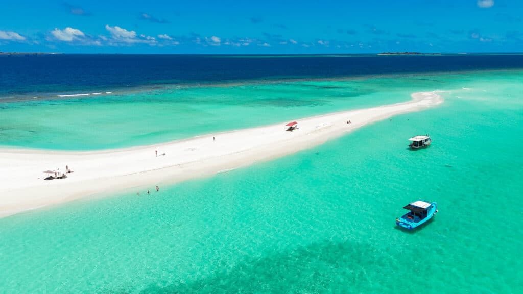 Long white sandbank stretching across turquoise and deep blue ocean with a few boats anchored nearby.
