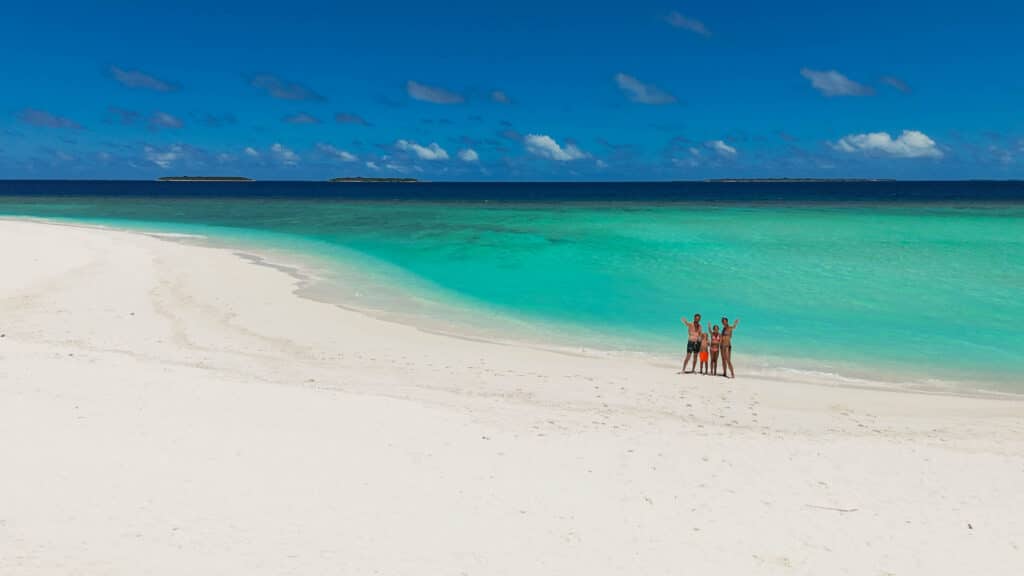 Family poses together on a wide white beach with turquoise sea at a sunny sandbank maldives.