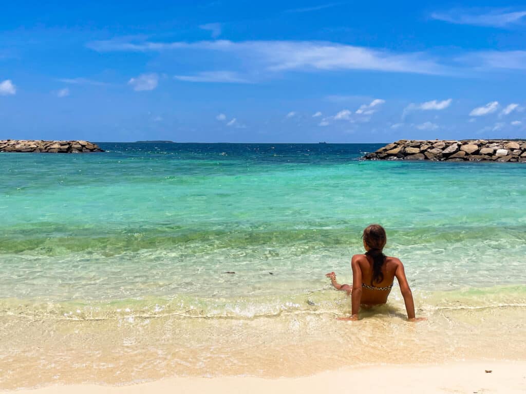 Girl sitting at the shoreline of a Maldivian beach looking out at the clear, shallow water on a sunny day.