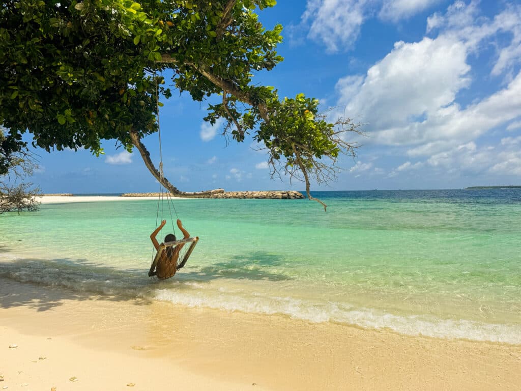 A guest swings over shallow turquoise water from a tree-hung seat on the shore at Kiha Beach.