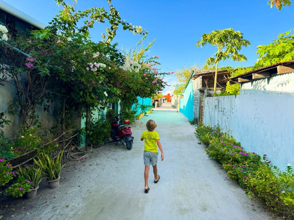 A child walks along a sandy village lane lined with flowers and colorful walls on the local island near Kiha Beach.