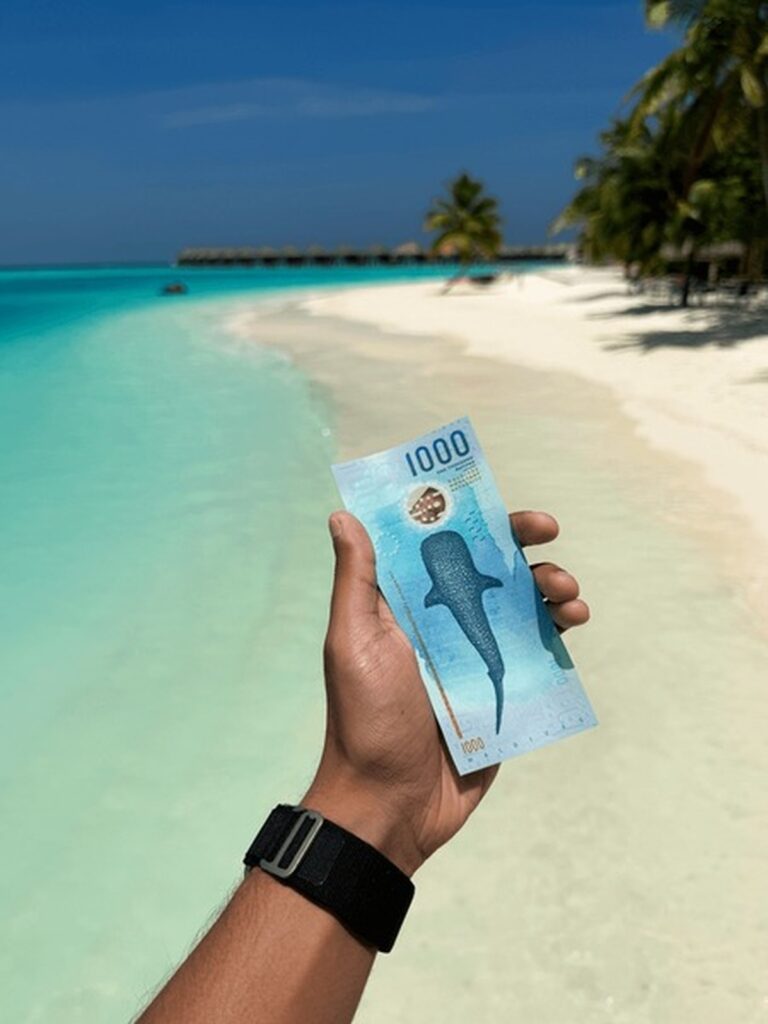 Mano sosteniendo un billete de 1000 rufiyaas maldivas en una playa de arena blanca.