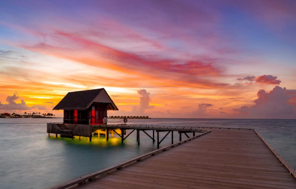 Los mejores resorts económicos en Maldivas: 10 opciones asequibles 2 embarcadero de llegada de madera que conduce a un pabellón sobre el agua al atardecer en un resort económico en Maldivas