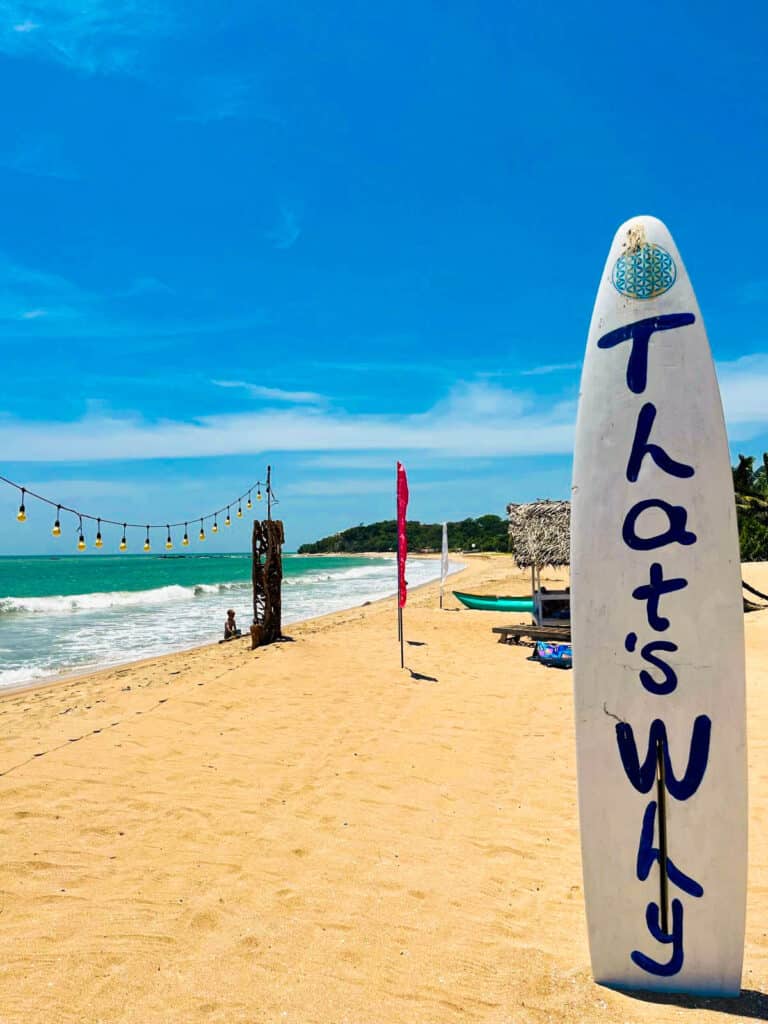 Sunny Sri Lankan beach with surfboard sign reading “That’s Why” and turquoise sea.