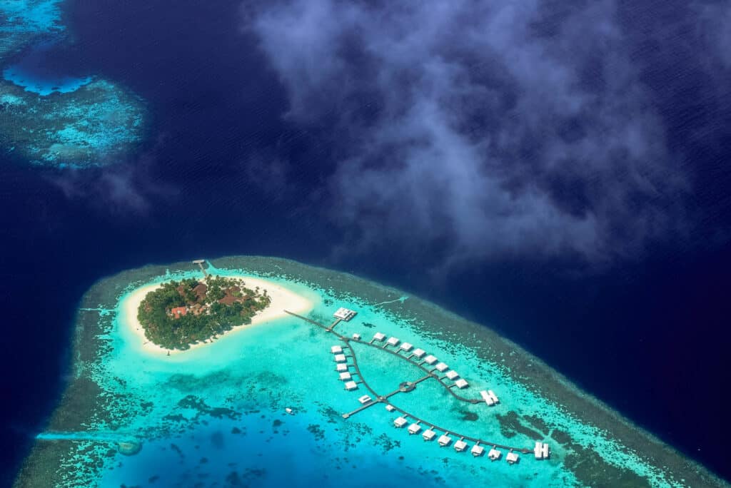 Aerial view of a small Maldivian resort island surrounded by bright turquoise lagoon and overwater villas.