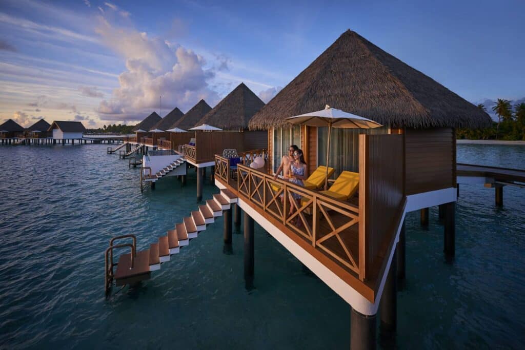 Los mejores resorts de Maldivas con mejor relación calidad-precio: 10 islas privadas que superan su precio 17 pareja de pie en la terraza de una villa sobre el agua con tumbonas amarillas al atardecer en Mercure Maldives Kooddoo