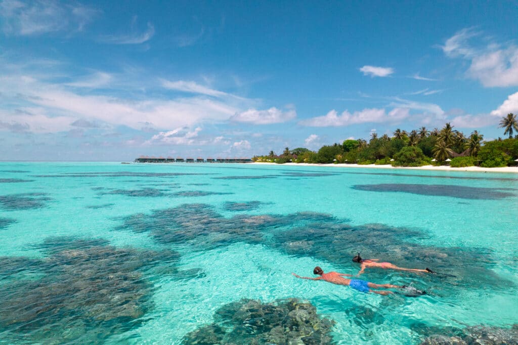 snorkeller over shallow coral reef at Komandoo Island Resort Maldives with overwater villas in background