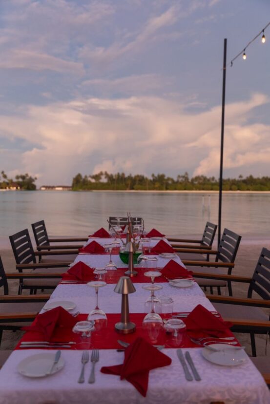 Dhigurah Maldives: The Best Local Island for Whale Shark Encounters 13 long dining table set with red napkins and glasses by the water at dusk overlooking a lagoon